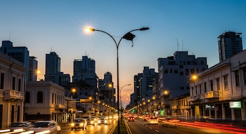 Calle de ciudad al atardecer con edificios, farolas encendidas y una apagada con parte rota, automóviles con luces y rastros de luz.