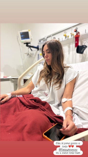 Floppy Tesouro recostada en una cama de hospital, con bata médica, vía intravenosa en el brazo izquierdo y una manta roja. Fondo de equipo médico