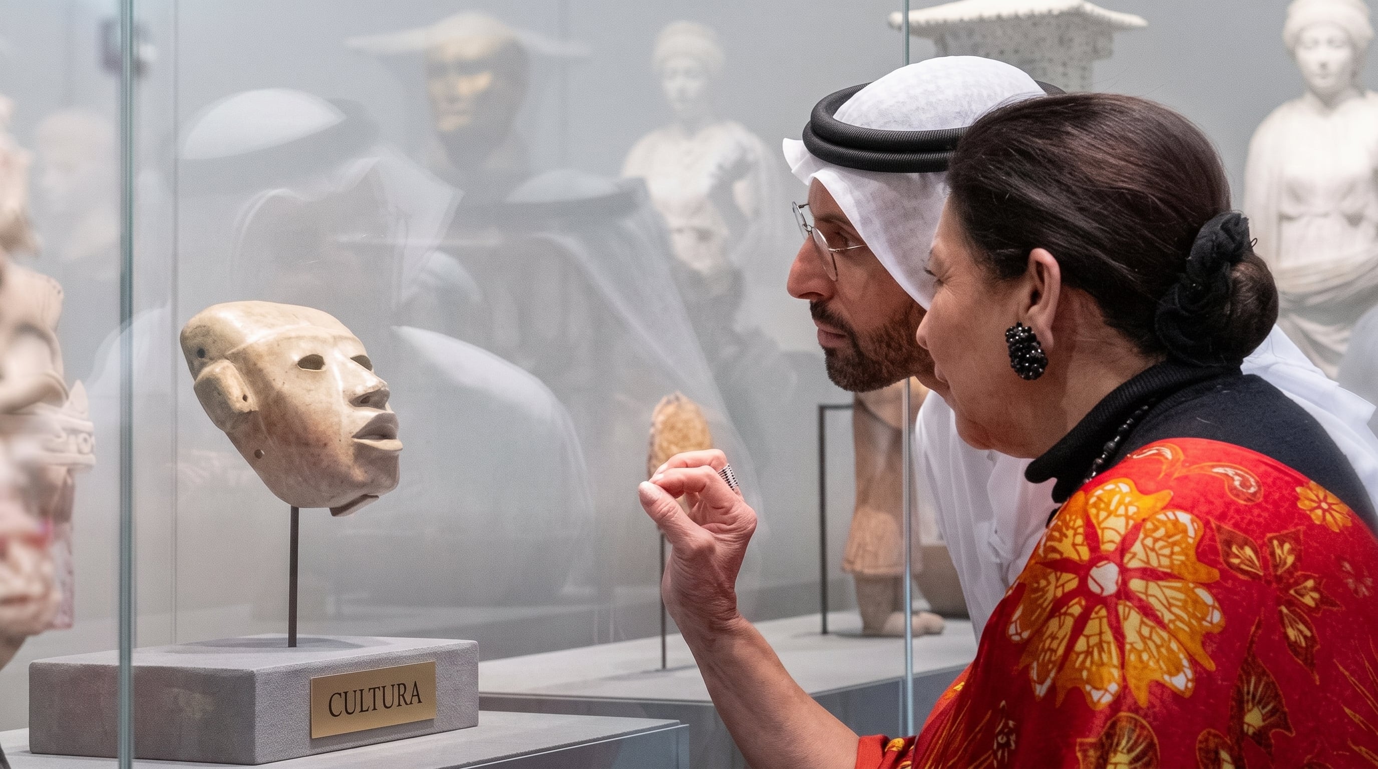 Un hombre con vestimenta tradicional árabe y una mujer observan una máscara de piedra expuesta dentro de una vitrina de museo. (Imagen Ilustrativa Infobae)