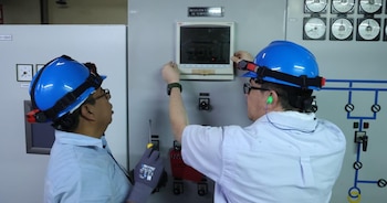 Dos técnicos con cascos de seguridad azules y lentes revisan un panel de control con medidores y una pantalla. Uno sostiene un destornillador