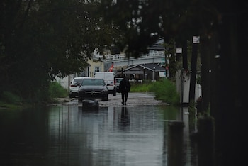 Vista frontal de una calle residencial inundada con agua gris reflejando el cielo, un hombre camina por el centro, y coches negros y blancos están a la derecha