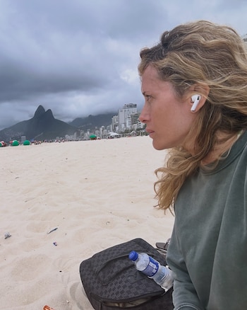 Una mujer con un auricular blanco permanece sentada en la arena junto a una mochila y una botella de agua, con montañas y edificios al fondo