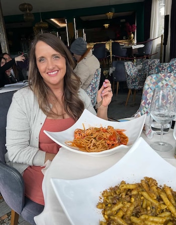 Una mujer con cabello castaño sonríe a la cámara en un restaurante, sosteniendo un tenedor sobre un plato de espagueti con langosta. Hay otro plato de pasta