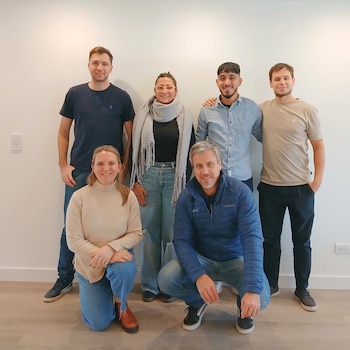 Seis personas en dos filas: tres hombres y una mujer de pie, y un hombre y una mujer arrodillados, sonriendo frente a una pared blanca con piso de madera