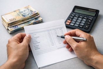 Manos sostienen un documento con datos y un bolígrafo. A la izquierda hay billetes de cien y cincuenta soles peruanos apilados, y una calculadora.