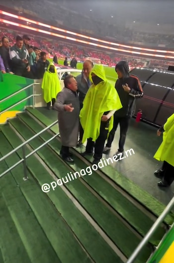 Miguel Herrera con abrigo gris gesticula junto a personal con impermeables amarillos en las escaleras de un estadio de fútbol
