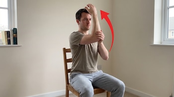 Un hombre sentado en una silla, con una camiseta y pantalones de chándal, realiza un ejercicio de hombro. Una flecha roja indica el movimiento del brazo.