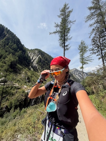 Una mujer con pañuelo rojo y chaleco deportivo se toma un autorretrato en un sendero rodeado de pinos y montañas al fondo