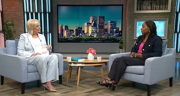 Dos mujeres sentadas en sillones celestes conversan en un estudio de televisión con una pantalla de fondo que muestra una ciudad