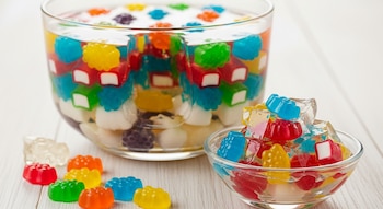 Recipiente de vidrio grande con gelatina y gomitas de colores. Cuenco pequeño con gelatina y gomitas. Varias gomitas sueltas sobre mesa blanca.