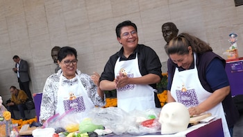 Tres personas con delantales blancos trabajan con masa de colores y una calavera de azúcar sobre una mesa