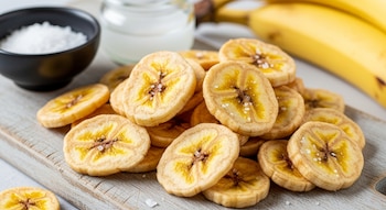 Primer plano de chips de banana crocantes y dorados con sal sobre una tabla de madera. Un cuenco de sal y bananas frescas son visibles al fondo.