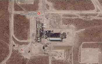 Vista aérea satelital de una planta industrial con tuberías y estructuras, ubicada en un terreno árido con caminos y un marcador rojo