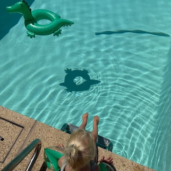 Pies de un niño rubio metidos en el agua de una piscina, al borde. Un flotador inflable verde con forma de cocodrilo flota
