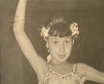 Primer plano de una niña con cabello oscuro, flequillo, diadema, flores en el cabello, pendientes y vestido