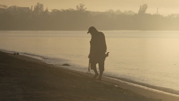 Silueta de un hombre con gorra y herramientas caminando descalzo por la orilla de una playa arenosa y brumosa al amanecer