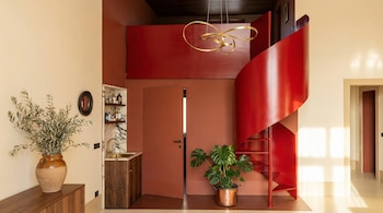 Interior moderno con una escalera caracol roja brillante que asciende en espiral, paredes terracota y beige, un bar con fregadero dorado, y una planta Monstera.