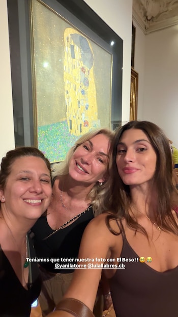 Tres mujeres sonríen a la cámara, posando frente a la famosa pintura 'El Beso' de Gustav Klimt, con tonos dorados predominantes. La obra está enmarcada