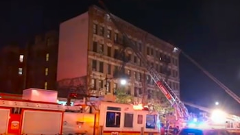 Vista nocturna de camiones de bomberos con luces intermitentes y escaleras extendidas hacia un edificio antiguo de varias plantas en Nueva York