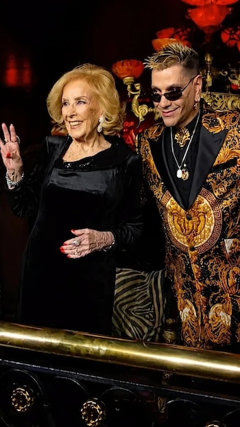 Mirtha Legrand, de cabello rubio, sonríe y saluda junto a Roberto Piazza, con gafas de sol y chaqueta dorada y negra, frente a una baranda