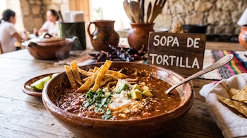 Plato de barro con caldo rojo, tiras de tortilla, aguacate, queso y cilantro, junto a un cartel de madera que dice SOPA DE TORTILLA.