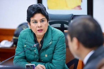 Mujer con cabello oscuro y chaqueta acolchada verde sentada en una mesa. Hay un micrófono y un teléfono móvil, y una persona borrosa de espaldas