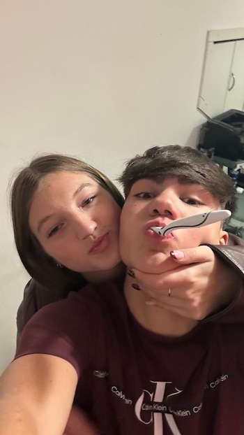 Mujer joven de cabello castaño abraza a hombre joven por el cuello. Él tiene un cubierto blanco en la boca y viste camiseta oscura. Fondo claro