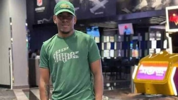 Hombre joven con gorra y camiseta verdes con logo de cocodrilo. Fondo desenfocado con pantallas electrónicas y una máquina amarilla y púrpura