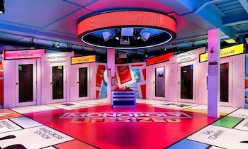 Interior de sala de juegos con el tablero de Monopoly gigante en el suelo rojo, paredes blancas con puertas y carteles de propiedades, e iluminación colorida