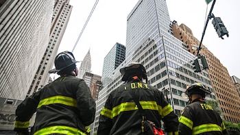 Tres bomberos con cascos y uniformes de espaldas, mirando hacia arriba a rascacielos, cables, un semáforo y la cúpula del edificio Chrysler