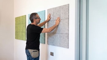 Un hombre de perfil con gafas instala un panel acústico gris en una pared blanca, junto a paneles verdes y azules. Viste camiseta negra y jeans.