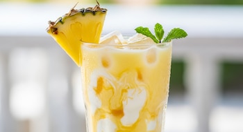 Primer plano de un vaso de agua de piña con leche condensada y hielo, decorado con una rodaja de piña y ramitas de menta fresca. El vaso está frío y húmedo.