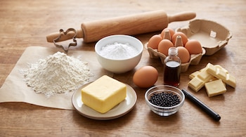 Harina, mantequilla, azúcar, huevos, chocolate blanco, esencia de vainilla, un rodillo y un cortador de galletas sobre una mesa de madera.