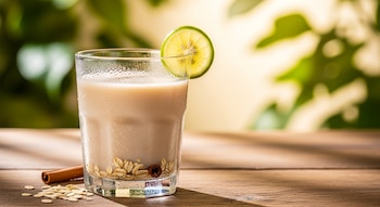 Vaso de agua de avena fría con gotas de condensación, decorado con una rodaja de lima, sobre una mesa de madera con avena y una rama de canela, fondo verde borroso.