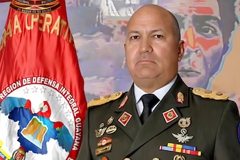 Hombre calvo con uniforme militar y medallas posa frente a bandera roja con texto dorado y emblema, y un rostro difuminado en el fondo
