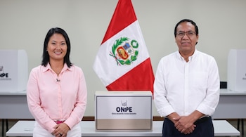 Una mujer y un hombre, candidatos presidenciales, posan sonrientes junto a un ánfora de la ONPE. Al fondo, la bandera de Perú y cabinas de votación.