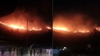 Vista nocturna dividida de una línea de fuego y humo en la ladera de una colina oscura sobre varias viviendas