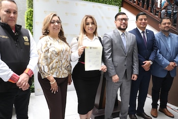 Greicy De Leon, con camisa blanca y falda negra, sonríe sosteniendo un documento. La acompañan cinco hombres en traje en un evento formal
