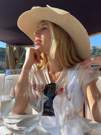 Maren Morris con cabello rubio, sombrero de paja y camiseta blanca con estampado, sentada en una terraza al aire libre bajo el sol, con gafas de sol colgadas en su camisa