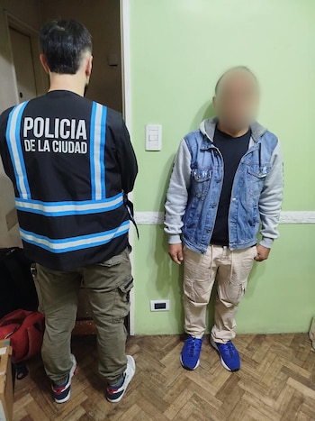 Un oficial de la Policía de la Ciudad, de espaldas, junto a un hombre con el rostro borroso, vestido con chaqueta vaquera y pantalones claros, en un interior