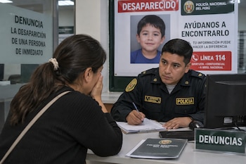 Un agente policial toma datos en un escritorio frente a una mujer, ante un afiche de un niño desaparecido y la señalización de denuncias