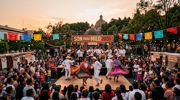 Parejas con trajes regionales bailan sobre una tarima rodeadas de público, frente a un escenario con músicos y puestos de artesanías.
