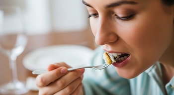 Primer plano de una mujer de piel morena comiendo. Sostiene un tenedor con un bocado y lo lleva a la boca, con los ojos semicerrados. En el fondo, mesa con platos y copas desenfocados.