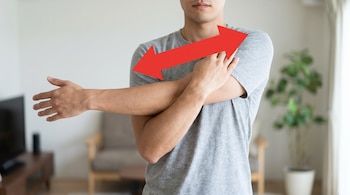 Primer plano de un hombre con camiseta gris realizando un estiramiento cruzado de brazo en su casa. Una flecha roja indica el movimiento del brazo y hombro.