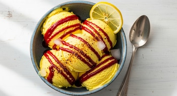 Primer plano de un tazón azul lleno de helado de limón amarillo con rayas de chamoy rojo, especias, cubos de hielo y una rodaja de limón. Una cuchara de metal descansa a su lado sobre una mesa blanca.