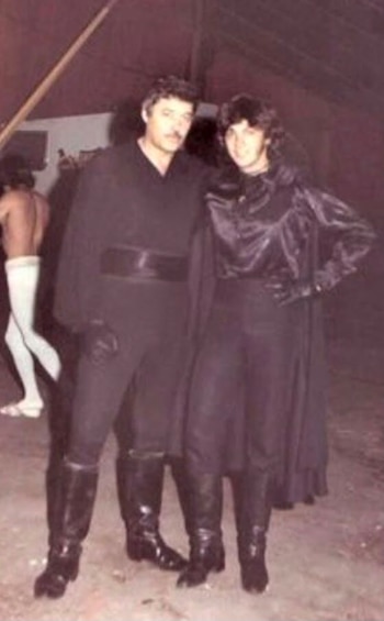Dos hombres vestidos con trajes negros y botas altas posan de pie en un escenario
