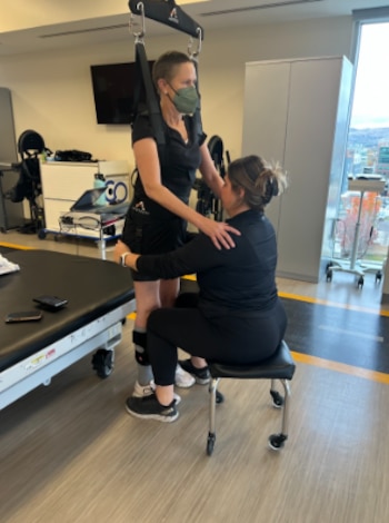 Una mujer con arnés de suspensión y mascarilla de pie, asistida por otra mujer sentada en un taburete. Sala con equipo médico, mesa y ventana al fondo