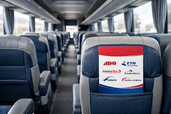 Interior de autobús con asientos azules y grises vacíos, pasillo central, ventanas y un folleto con logotipos de empresas de transporte en el respaldo de un asiento.
