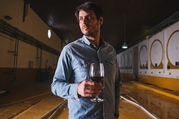 Un hombre de mediana edad con camisa de mezclilla sostiene una copa de vino tinto, mirando hacia un lado en una bodega con grandes tanques blancos y amarillos al fondo