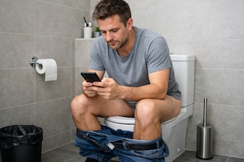 Hombre caucásico con camiseta gris y pantalones vaqueros bajados sentado en un inodoro blanco y usando un teléfono móvil en un baño con azulejos grises.